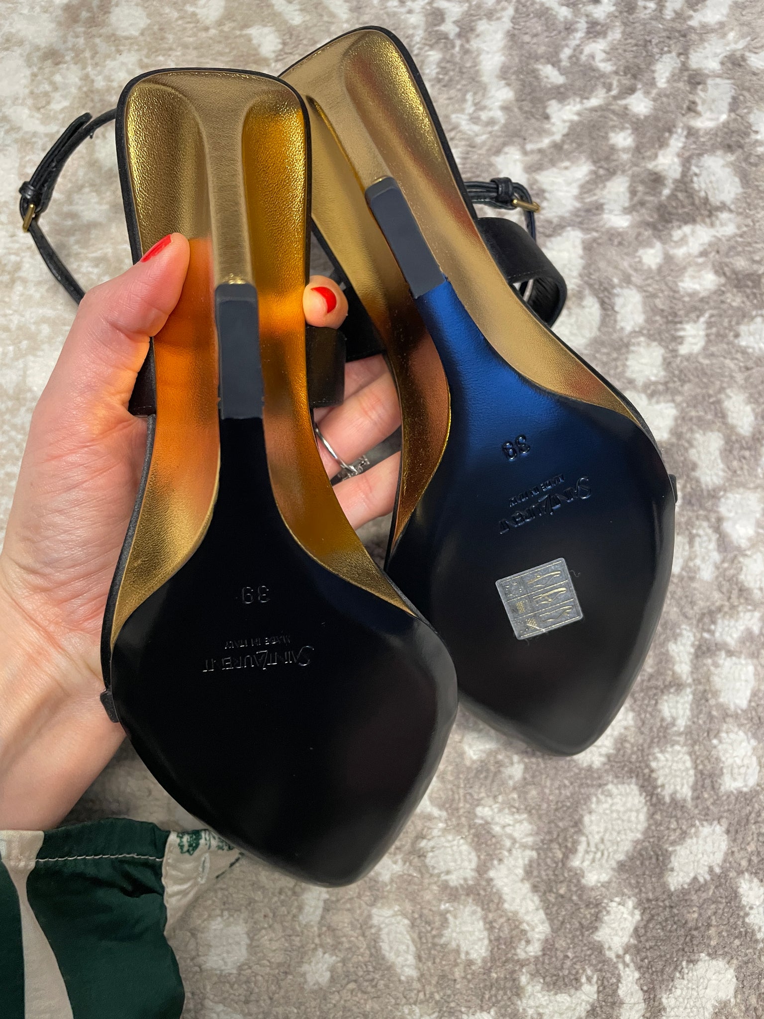 Black high-heeled shoes with gold soles held by a hand on a textured surface